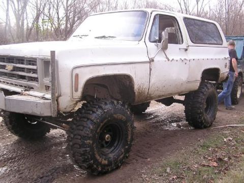 White SUV with lifted suspension and large off-road tires, mud splattered on body. Front grille visible