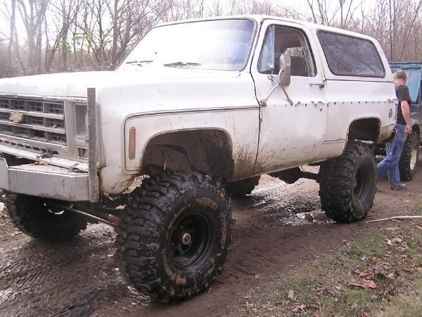 White SUV with lifted suspension and large off-road tires, mud splattered on body. Front grille visible