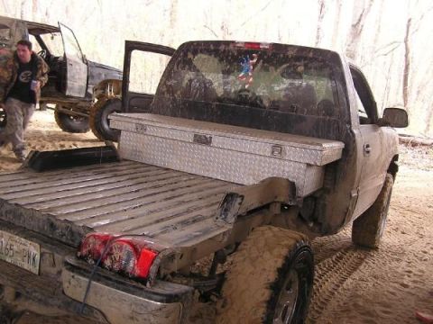 Rear view of a dirty pickup truck with a toolbox in the bed. Another vehicle is visible to the left