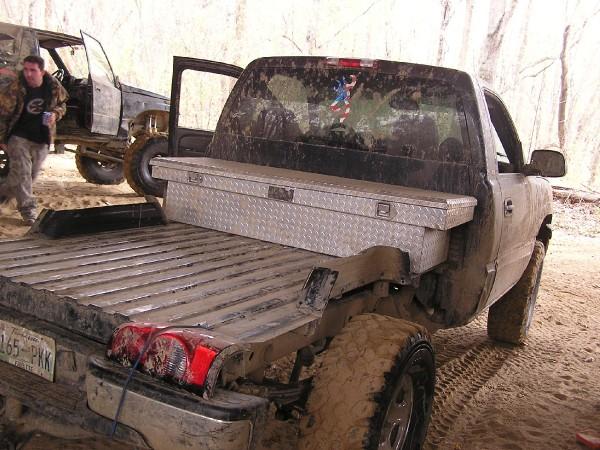 Rear view of a dirty pickup truck with a toolbox in the bed. Another vehicle is visible to the left