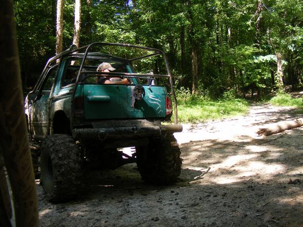 Green off-road vehicle with a roll cage, driving on a dirt path in the woods