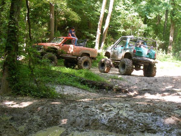 Orange off-road truck with a person on top, and a green off-road SUV on a muddy trail