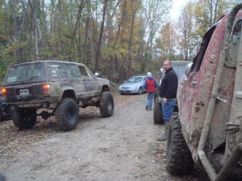 Muddy lifted Jeep Cherokee from rear, with other off-road vehicles and people
