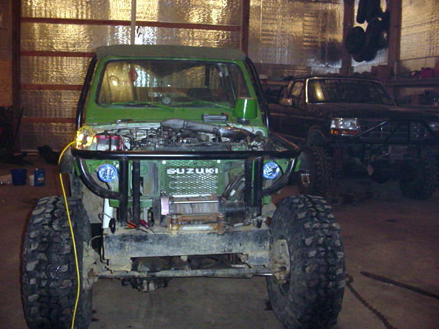 Green Suzuki Samurai with aftermarket bumper and winch, hood open, in a garage