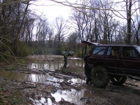 Several off-road vehicles stuck in mud and puddles in a wooded area. One maroon SUV with large tires is promin
