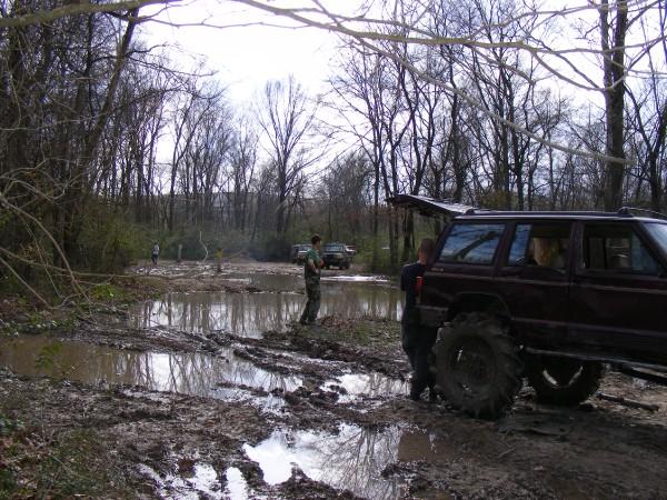 Several off-road vehicles stuck in mud and puddles in a wooded area. One maroon SUV with large tires is prominent