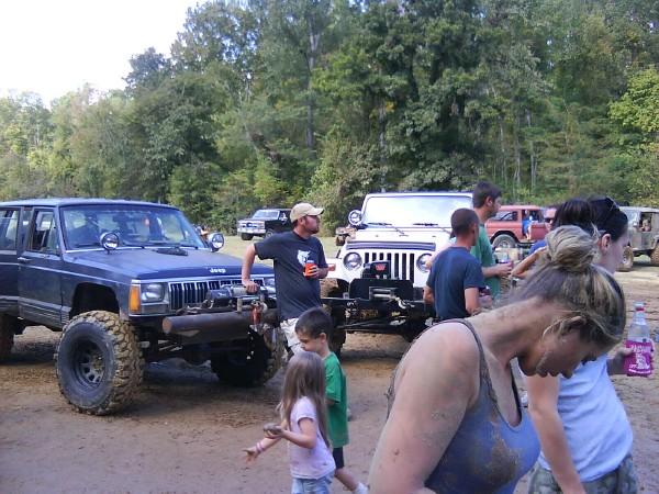 Blue Jeep Cherokee XJ with winch, white Jeep Wrangler, and other off-roaders gathered on a dirt path
