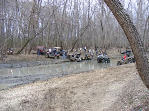 Multiple off-road vehicles navigating a muddy creek surrounded by trees