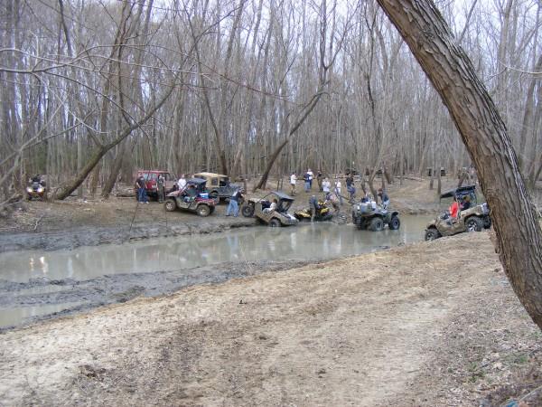 Multiple off-road vehicles navigating a muddy creek surrounded by trees