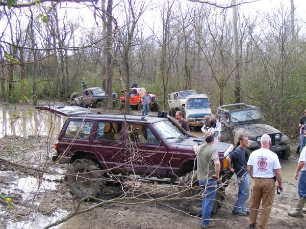 Several off-road vehicles stuck in mud and trees. A maroon Jeep Cherokee is in the foreground