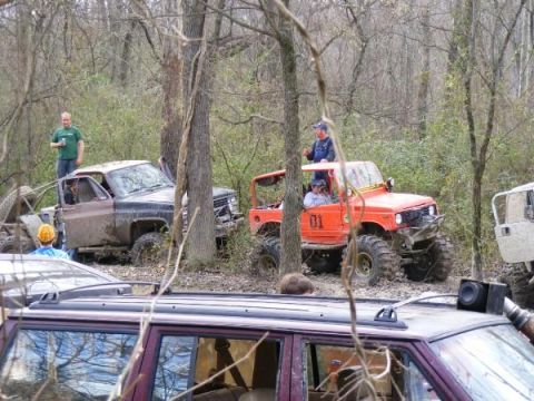 Orange off-road vehicle with number 01 on door, muddy truck, and other vehicles in woods