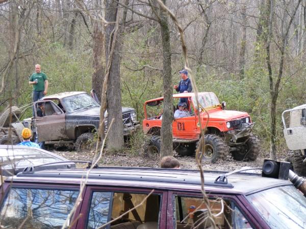 Orange off-road vehicle with number 01 on door, muddy truck, and other vehicles in woods