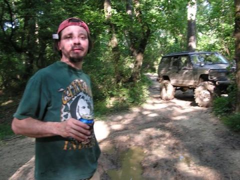 Man in red hat and green t-shirt holding a beer can, standing on a dirt path. A lifted, muddy off-road SUV is