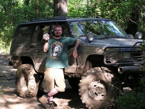 Man with can next to a very muddy SUV with large tires in a wooded area