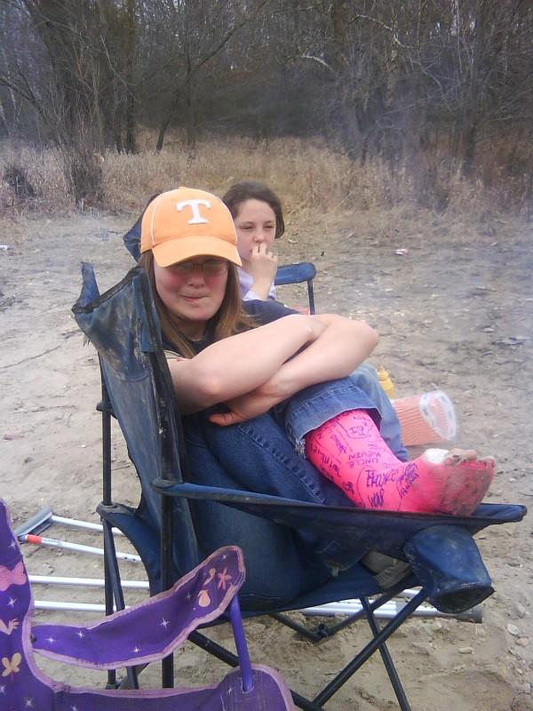 Young woman in orange cap and jeans sits with arms crossed, pink cast on her leg. Another girl sits behind her