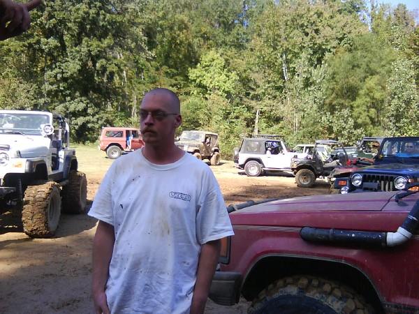 Man in white t-shirt with glasses, standing in a dirt lot with several off-road vehicles and trees in the background. A Supermotors logo is visible on a red vehicle in the foreground