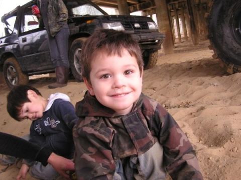 Two young boys in camouflage jackets on sandy ground with a black SUV behind them