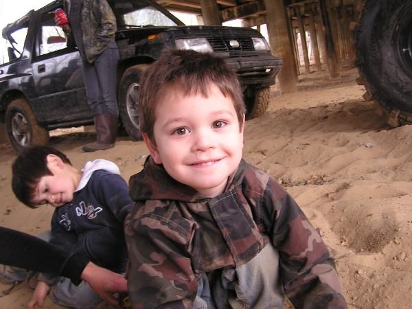 Two young boys in camouflage jackets on sandy ground with a black SUV behind them