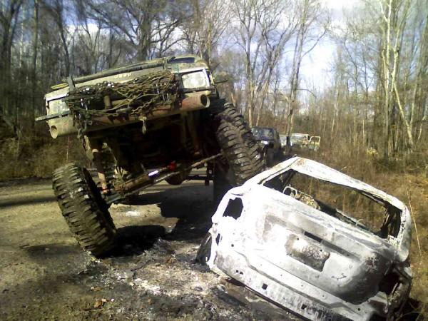 1978 Ford Bronco modified for off-roading, tilted on its side next to a burnt-out SUV