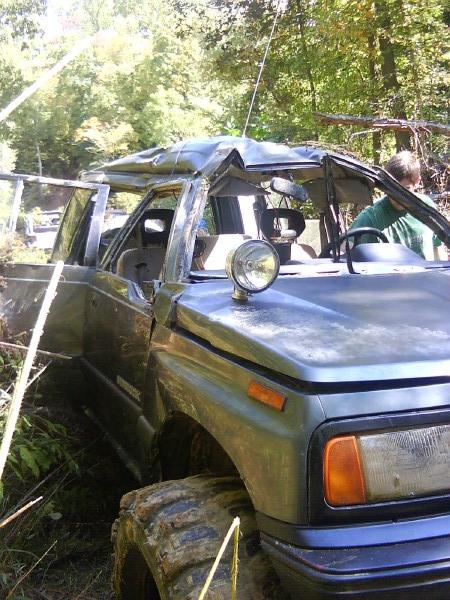 Damaged SUV with dented roof and open doors, stuck in wooded area. Large off-road tires are visible