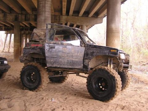 Muddy, lifted SUV with large tires parked under a bridge. Another vehicle is partially visible to the left