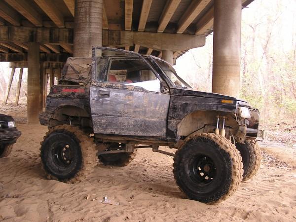 Muddy, lifted SUV with large tires parked under a bridge. Another vehicle is partially visible to the left
