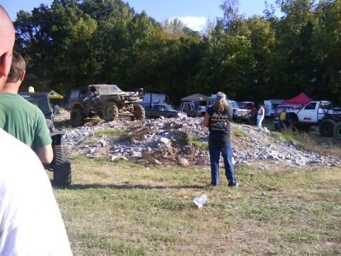 Several off-road vehicles are parked on a rocky incline with trees in the background. People are standing arou