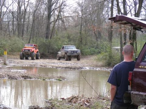 Two off-road vehicles are stuck in mud near trees. A person stands next to a maroon vehicle in the foreground