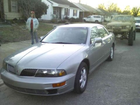 Silver sedan parked on asphalt, front quarter view. A person stands behind it. A lifted off-road vehicle is in