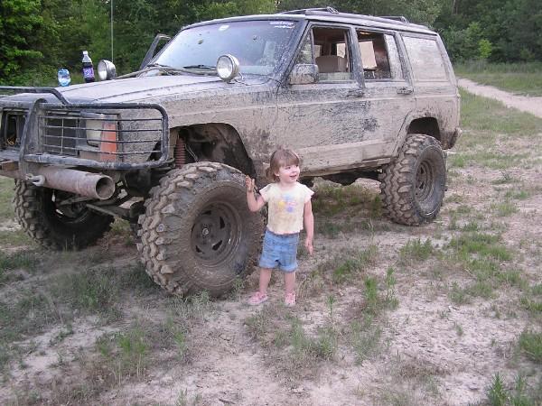 Muddy Jeep Cherokee XJ with large tires and a brush guard, a young child stands next to it