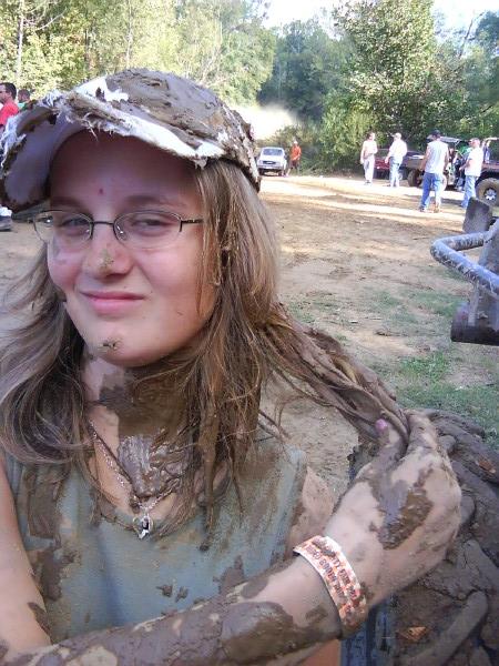 muddy person in foreground, several vehicles in background at an outdoor event