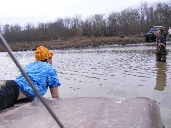 Person on vehicle hood in water with off-road vehicles in background