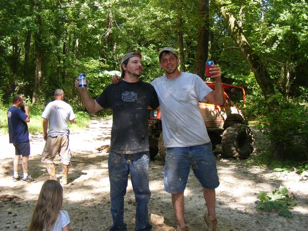 Two men holding beer cans in a muddy wooded area with an off-road vehicle behind them
