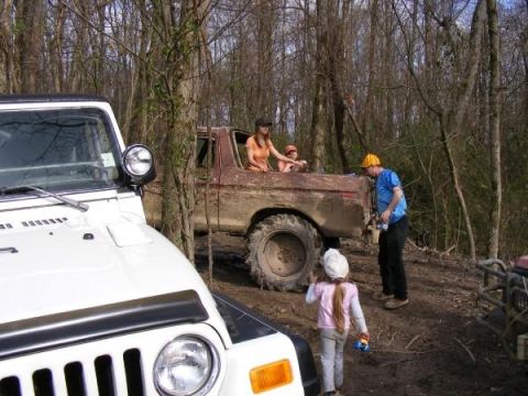 White Jeep in foreground, brown pickup truck with people in back, in wooded area