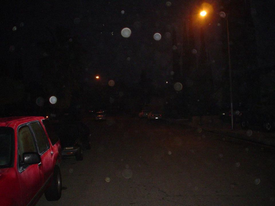 Night street scene with a red vehicle parked at the curb under orange streetlights, lens spots visible.