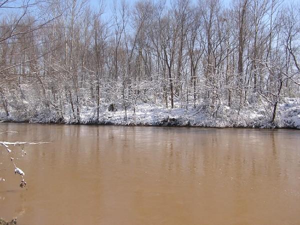 snowy trees line a muddy river under a clear sky. Supermotors.net logo in lower right
