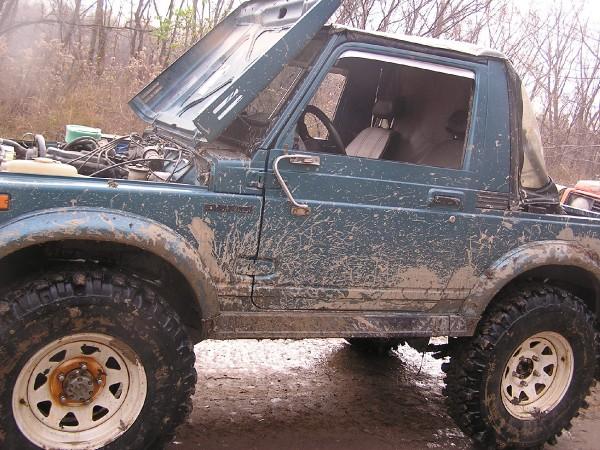Mud-covered off-road vehicle, hood open, showing engine details. Soft top is partially deployed