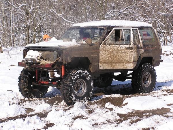 Mud-covered SUV with open driver's door stuck in snow, front grille removed