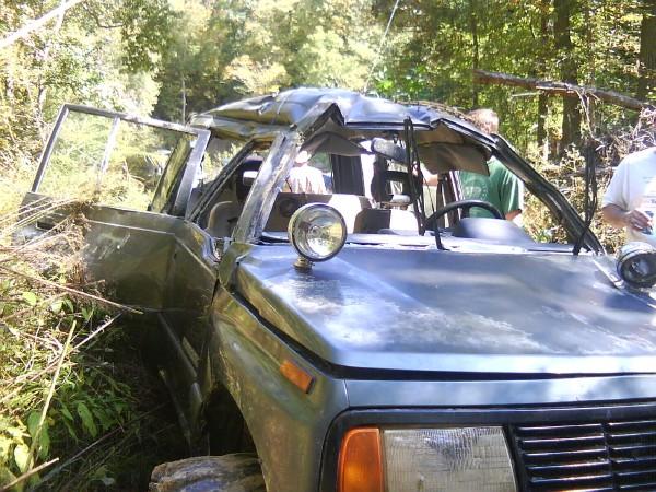 Damaged gray off-road vehicle with open doors and crumpled roof, partially obscured by foliage