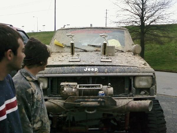 Muddy Jeep with two trophies on the hood, winch on front bumper, two people looking at it
