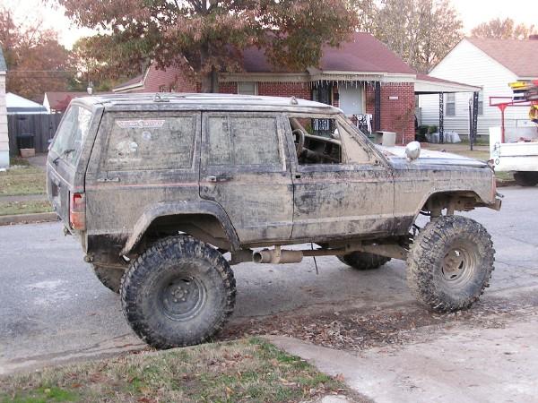 Mud-covered Jeep Cherokee XJ with oversized tires parked on a residential street