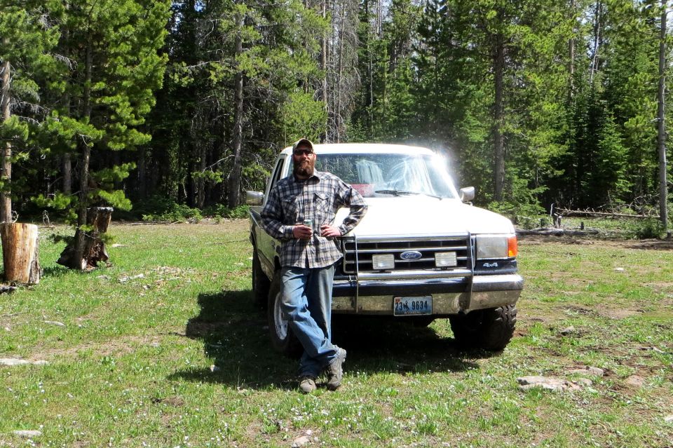 Guy leaning on a white Ford F-Series pickup, looks like late 80s model. It's parked in a grassy, wooded area
