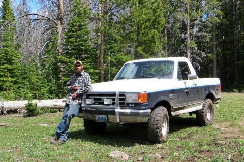 1989 Ford Bronco - A man leans against a white and dark blue Ford F-Series truck in a wooded area.