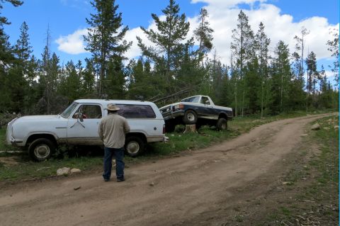 1989 Ford Bronco - Two trucks are parked on a dirt road surrounded by trees under a cloudy sky.