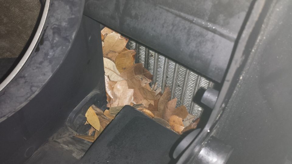 Dead brown leaves clogging a vehicle's cabin air intake and metal fins behind gray plastic housing.