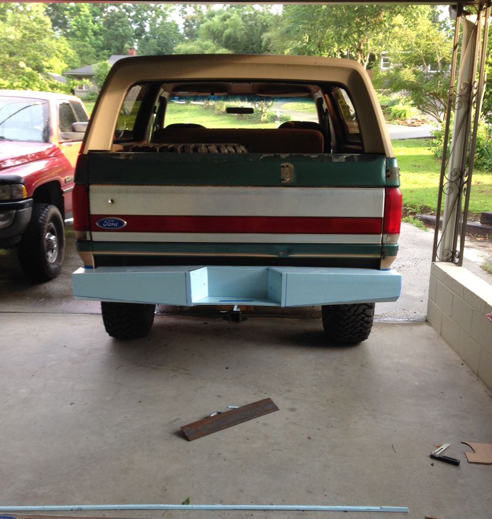 Rear view of a Ford Bronco with a light blue custom bumper. A red pickup truck is parked to the left