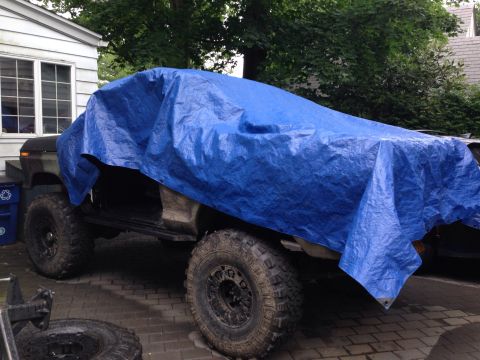1979 Ford Bronco - A vehicle, possibly a Ford Bronco, is covered by a blue tarp, with its large off-road tires visible.