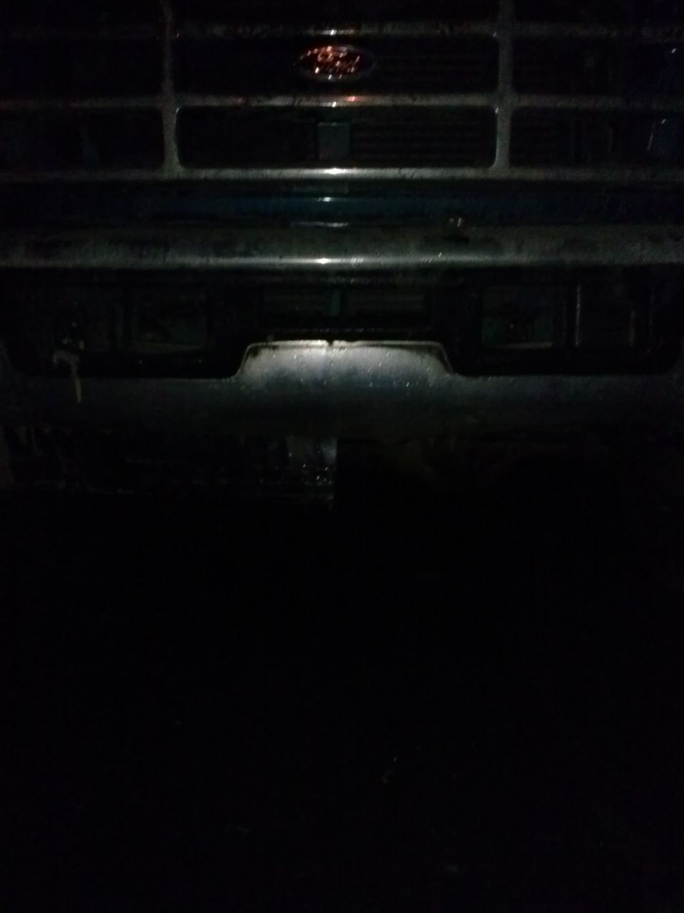 Dark front view of a 1990s Ford truck grille with a red Ford emblem. The bumper is visible
