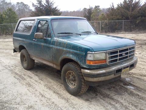 1996 Ford Bronco - Mud-covered 1996 Ford Bronco sits on a dirt track with trees in the background.
