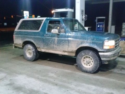 1996 Ford Bronco - A dirty teal Ford Bronco with a white top is parked at a gas station at night.